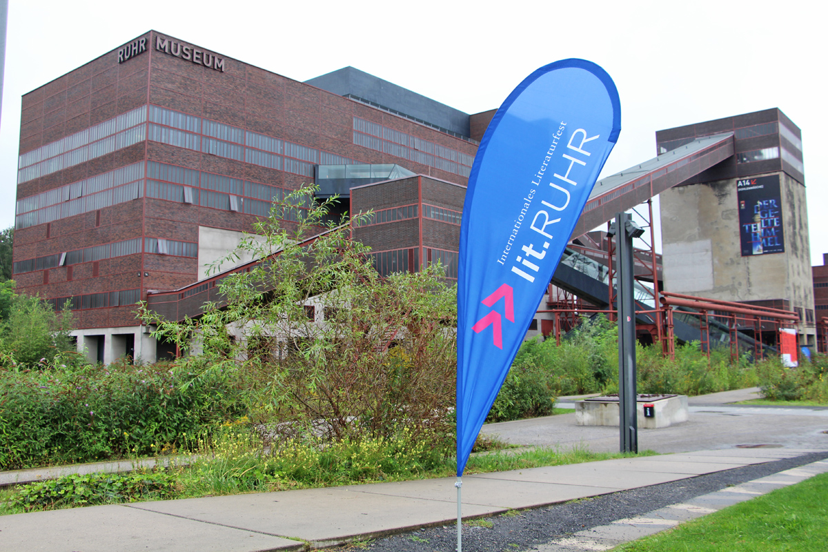 Das Bild zeigt das Logo der Lit.Ruhr vor einem Zollverein Gebäude