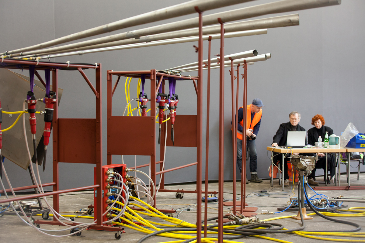 Das Bild zeigt eine Installation aus Abbauhämmern und drei Menschen im Hintergrund