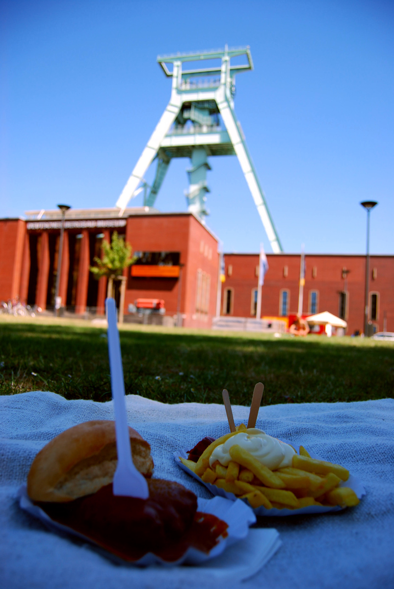 Das Foto zeigt Currywurst-Pommes vor der Kulisse des Deutschen Bergbaumuseums in Bochum