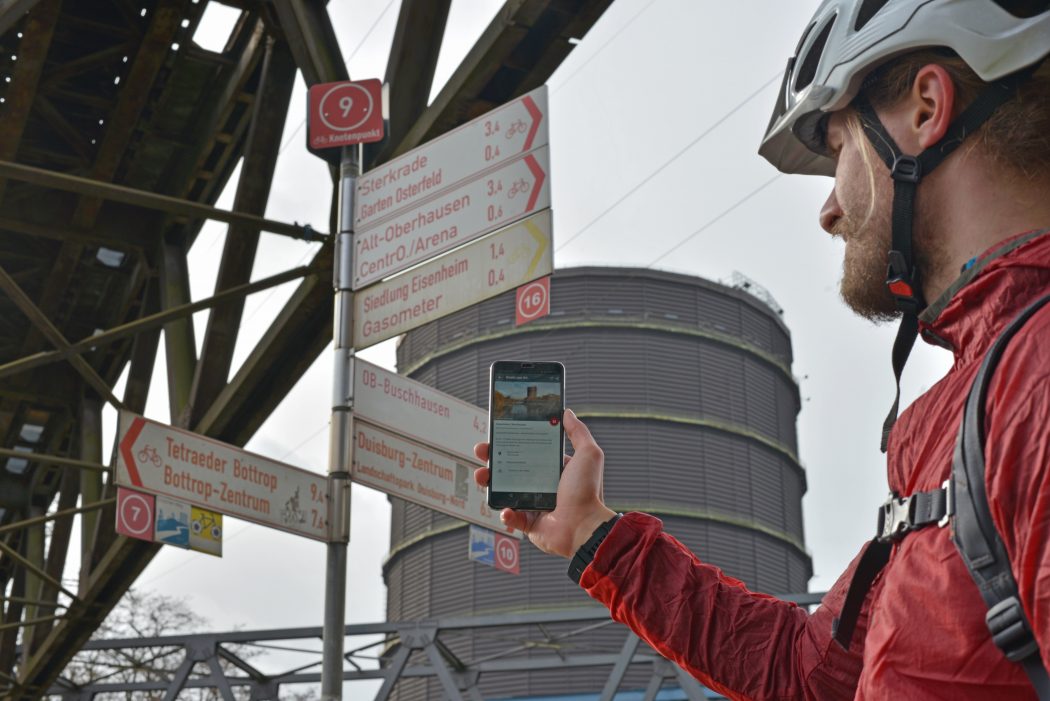 Das Foto zeigt einen Radfahrer vorm Gasometer Oberhausen, der den digitalen radtourenplaner.ruhr auf seinem Handy bedient