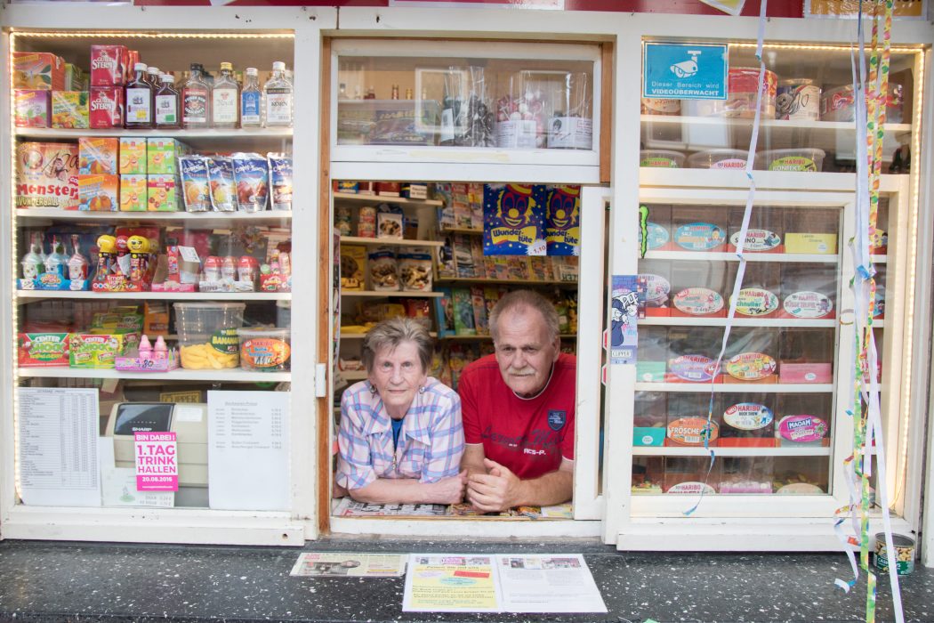 Eine ältere Frau und ein älterer Mann lehnen an der Theke eines kleinen Schaufensters, umgeben von Regalen, die mit bunten Snacks, Süßigkeiten und verschiedenen verpackten Waren gefüllt sind - eine Szene ganz typisch Ruhrpott. An der rechten Seite des Schaufensters hängen Luftschlangen.