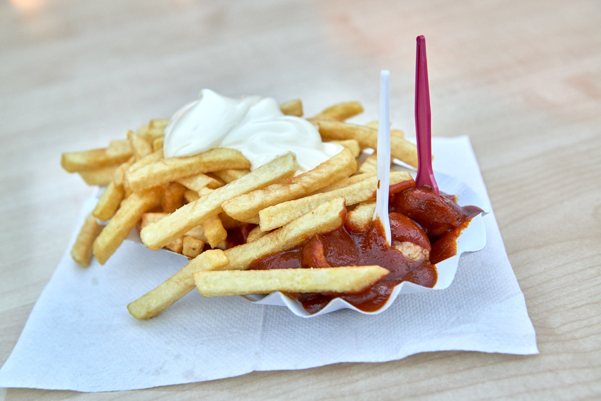 Eine Portion Pommes frites mit Mayonnaise und Wurstscheiben mit Curryketchup, präsentiert auf einem weißen Papiertablett mit zwei Plastikgabeln, die auf einer Serviette auf einem Holztisch liegen.