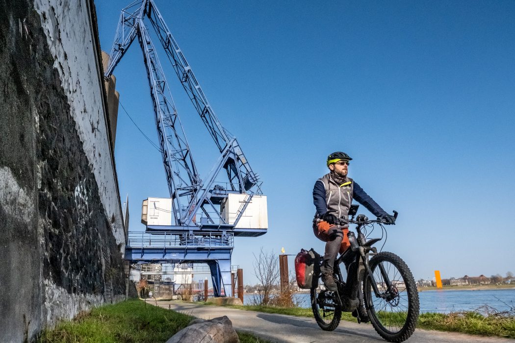 Das Foto zeigt einen Radfahrer am Rhein - hinter ihm ein Kran und die Rheinorange in Duisburg