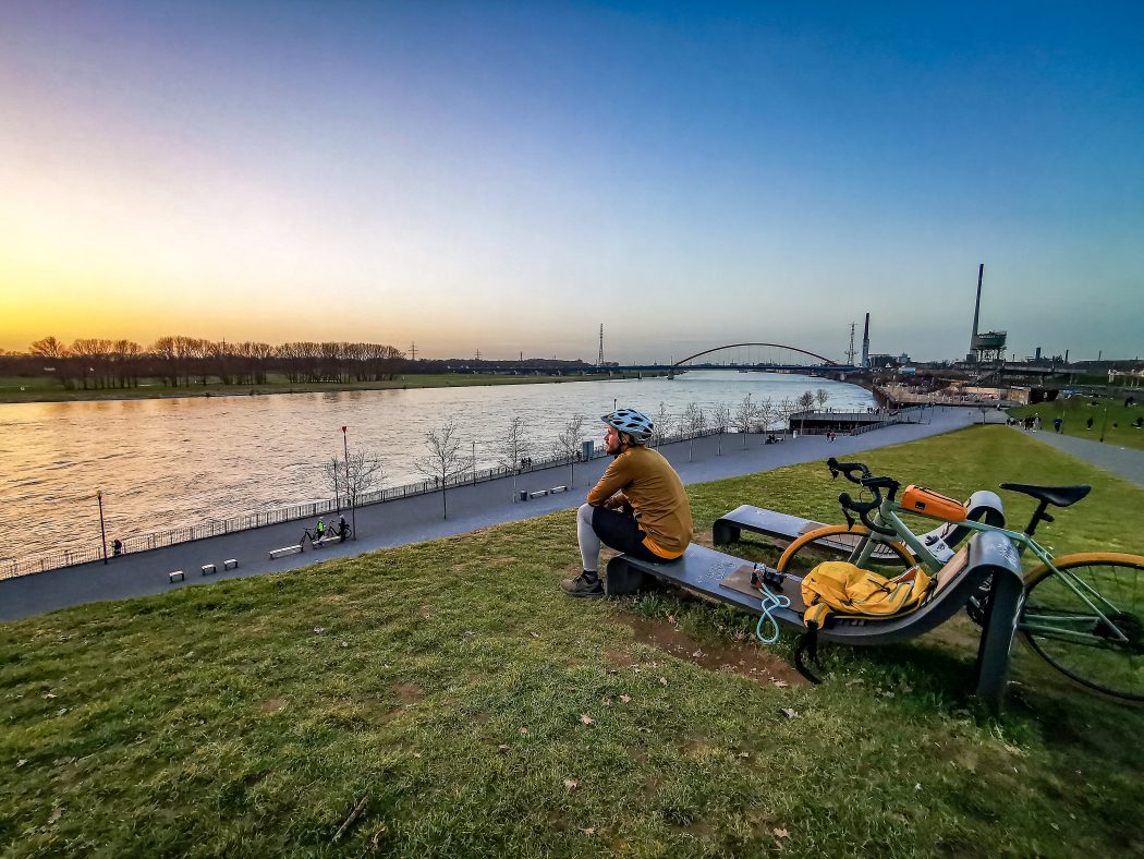 Das Foto zeigt einen Radfahrer mit Blick auf den Sonnenuntergang am Rhein in Duisburg