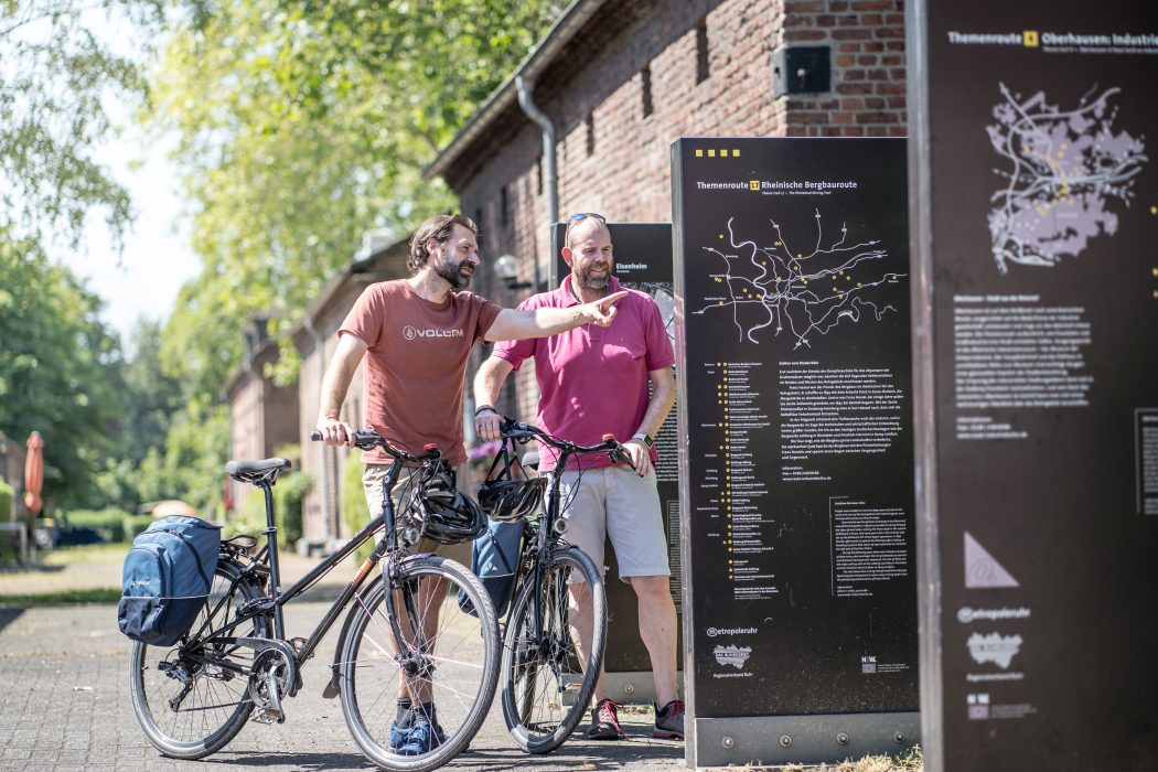 Das Foto zeigt zwei Radfahrer an den Info-Brammen der Route Industriekultur an der Arbeitersiedlung Eisenheim in Oberhausen