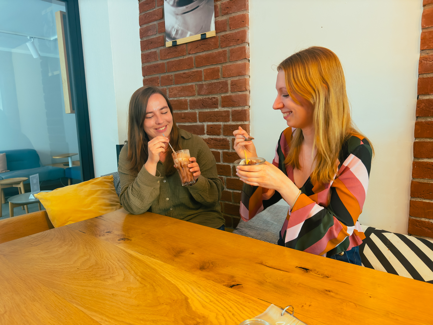 Zwei Frauen sitzen an einem Holztisch in einem gemütlichen Café, lächeln und genießen ihre Desserts. Die eine hält ein Dessertglas, die andere eine Schale. Sie scheinen ein freundliches Gespräch zu führen. Backsteinmauer und Sitzkissen im Hintergrund.