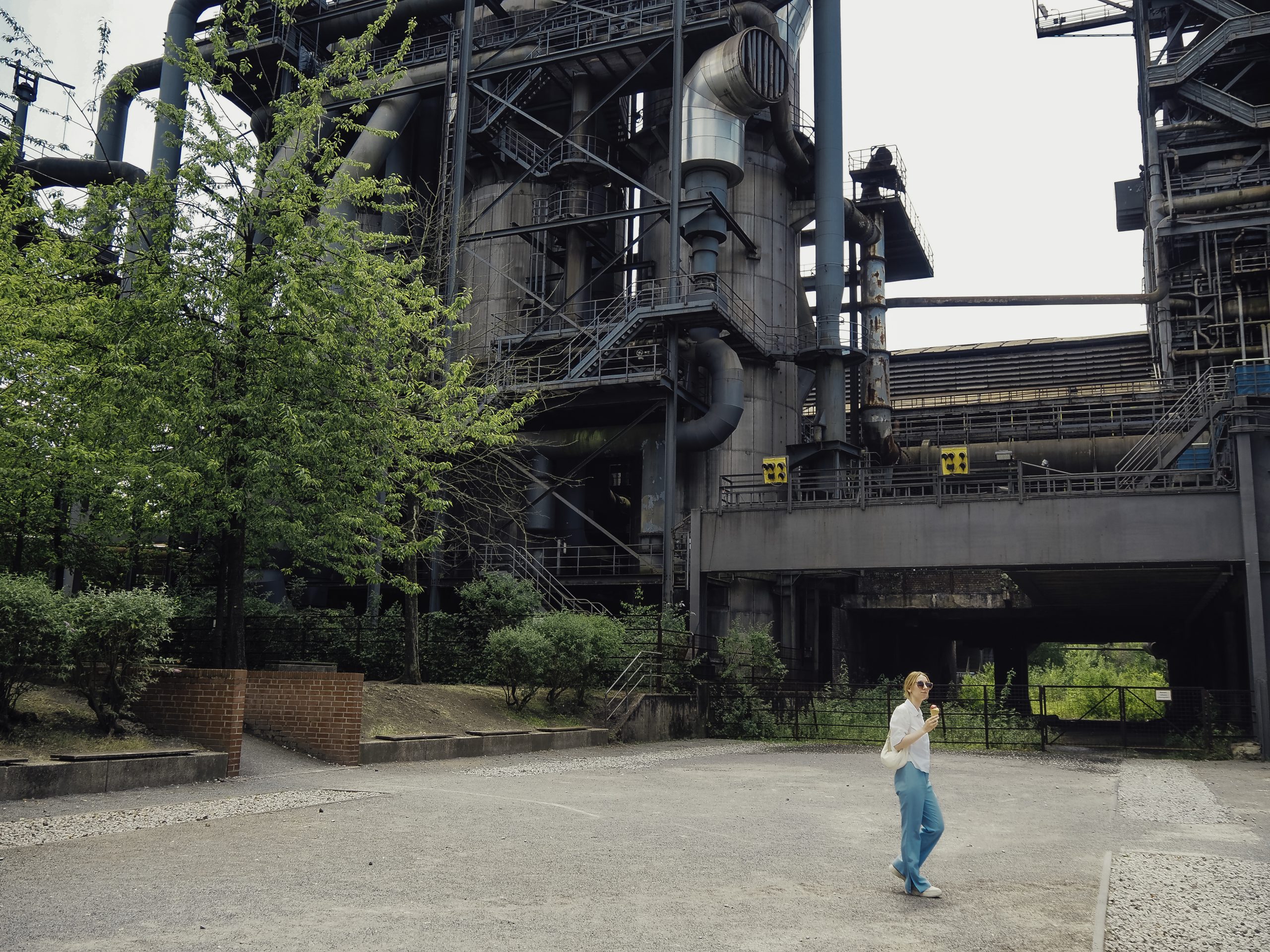 Eine Person, die ein weißes Hemd und eine blaue Jeans trägt, geht über eine offene, gepflasterte Fläche in der Nähe großer, industrieller Metallstrukturen, die von Grünflächen und Bäumen umgeben sind.