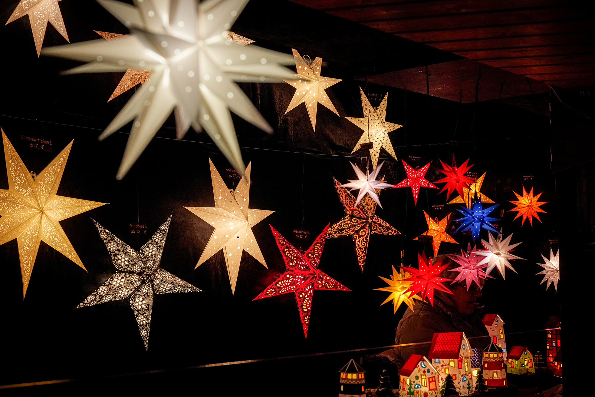 Bunte, beleuchtete sternförmige Laternen hängen vor einem dunklen Hintergrund und schaffen eine festliche Atmosphäre. Am unteren Rand des Bildes sind kleine, dekorative Modellhäuser zu sehen.