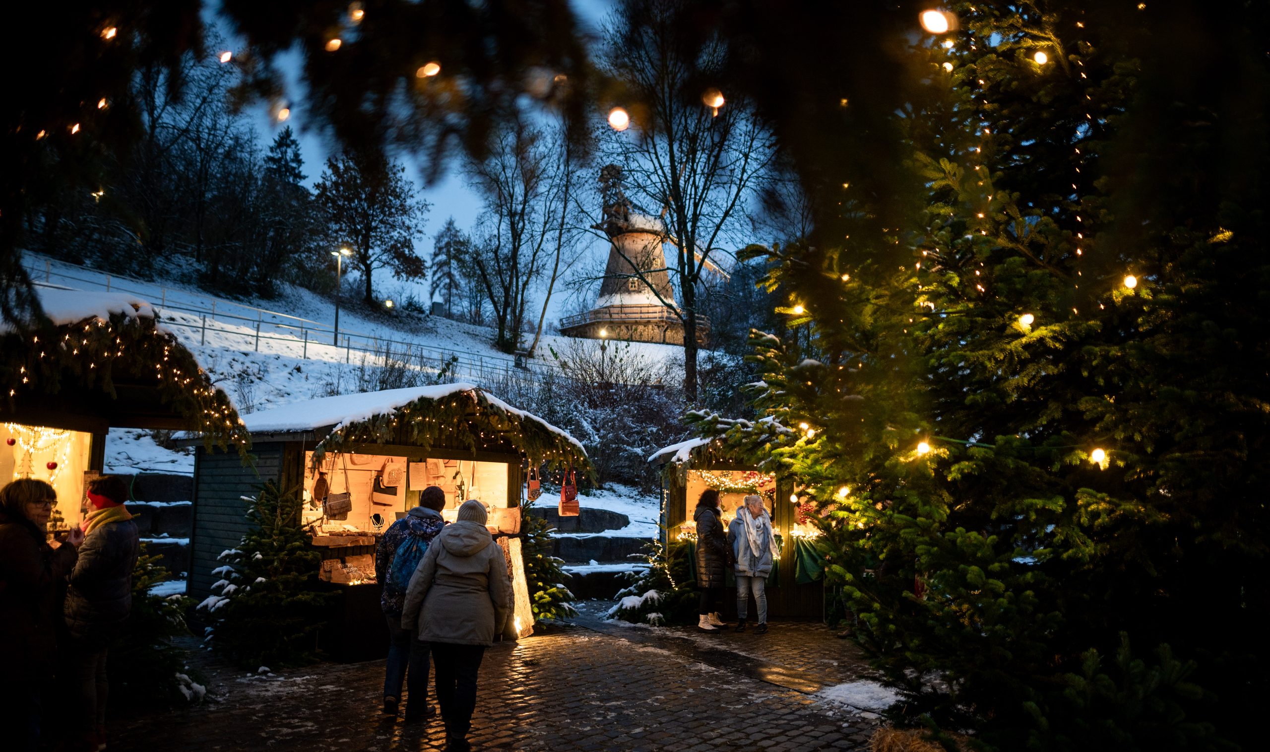 Ein gemütlicher Weihnachtsmarkt unter freiem Himmel in der Abenddämmerung, mit leuchtenden Lichterketten, geschmückten immergrünen Bäumen und Menschen, die an festlichen Ständen stöbern, umgeben von Schnee und bewaldeten Hügeln.