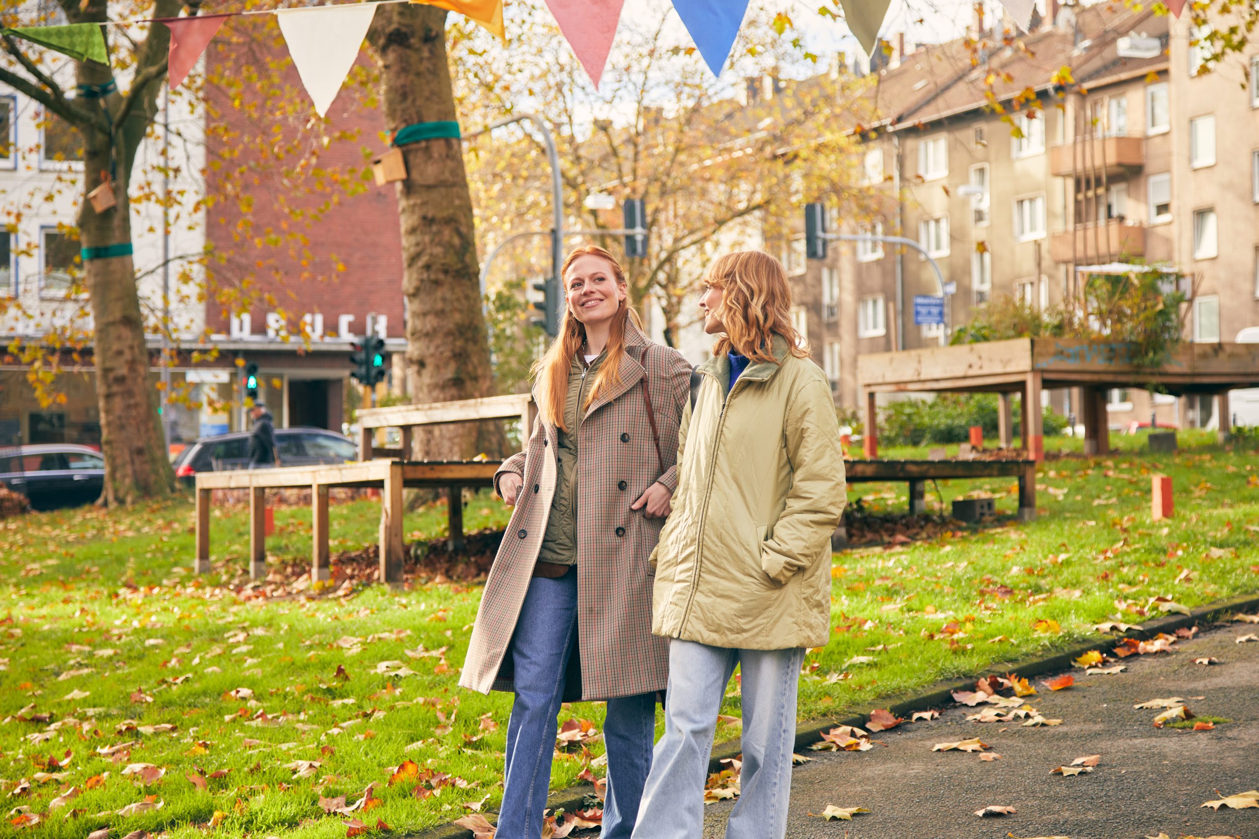 Zwei Frauen gehen an einem sonnigen Herbsttag zusammen in einem Park spazieren, lächeln und unterhalten sich. Über ihnen hängen bunte, dreieckige Wimpel, und das Gras ist mit abgefallenen Blättern bedeckt. Im Hintergrund sind Gebäude und Bäume zu sehen.