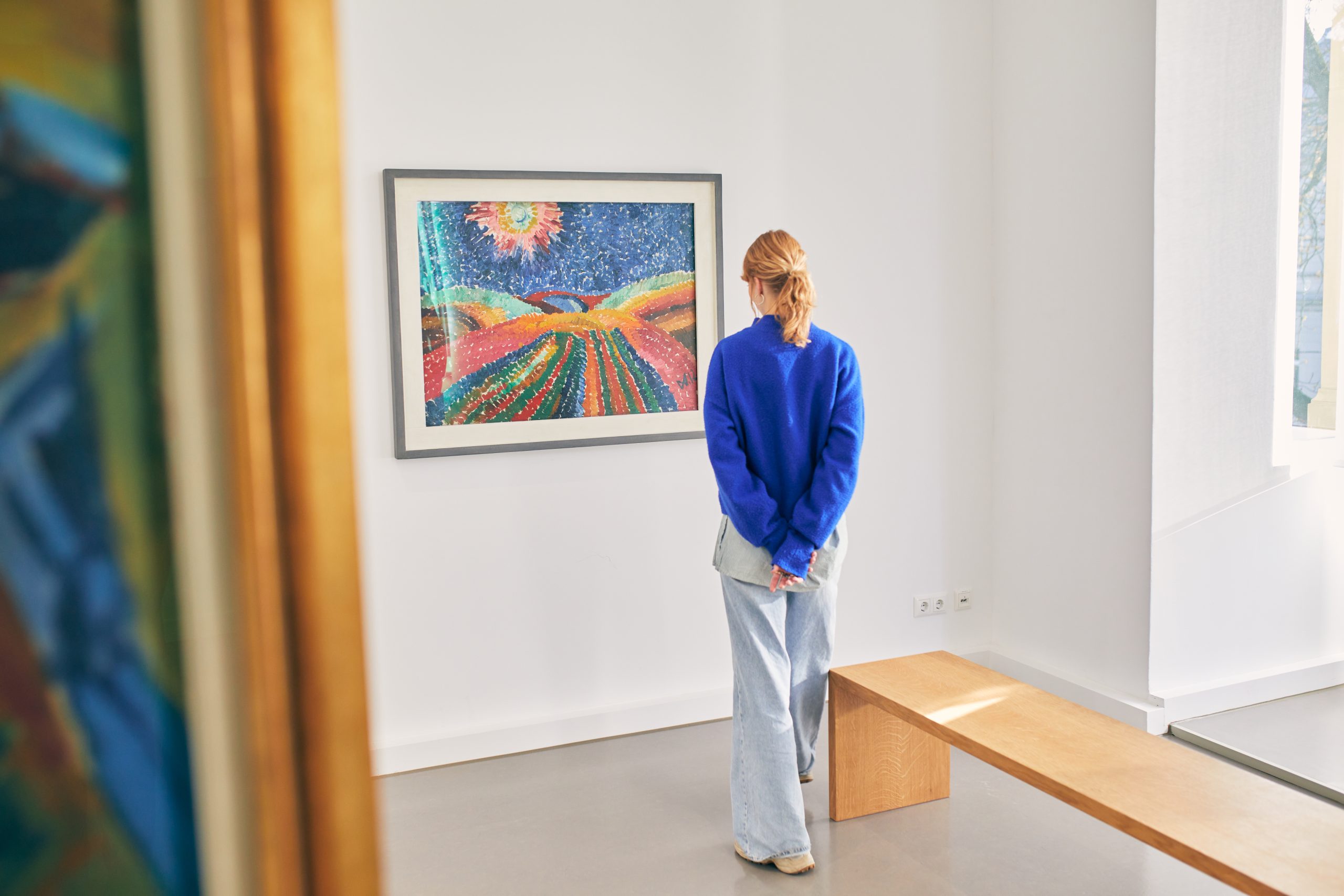 Eine Person in blauem Pullover und hellen Jeans steht in einer Kunstgalerie und betrachtet ein farbenfrohes, abstraktes Landschaftsgemälde an der Wand; durch ein nahe gelegenes Fenster strömt Sonnenlicht.