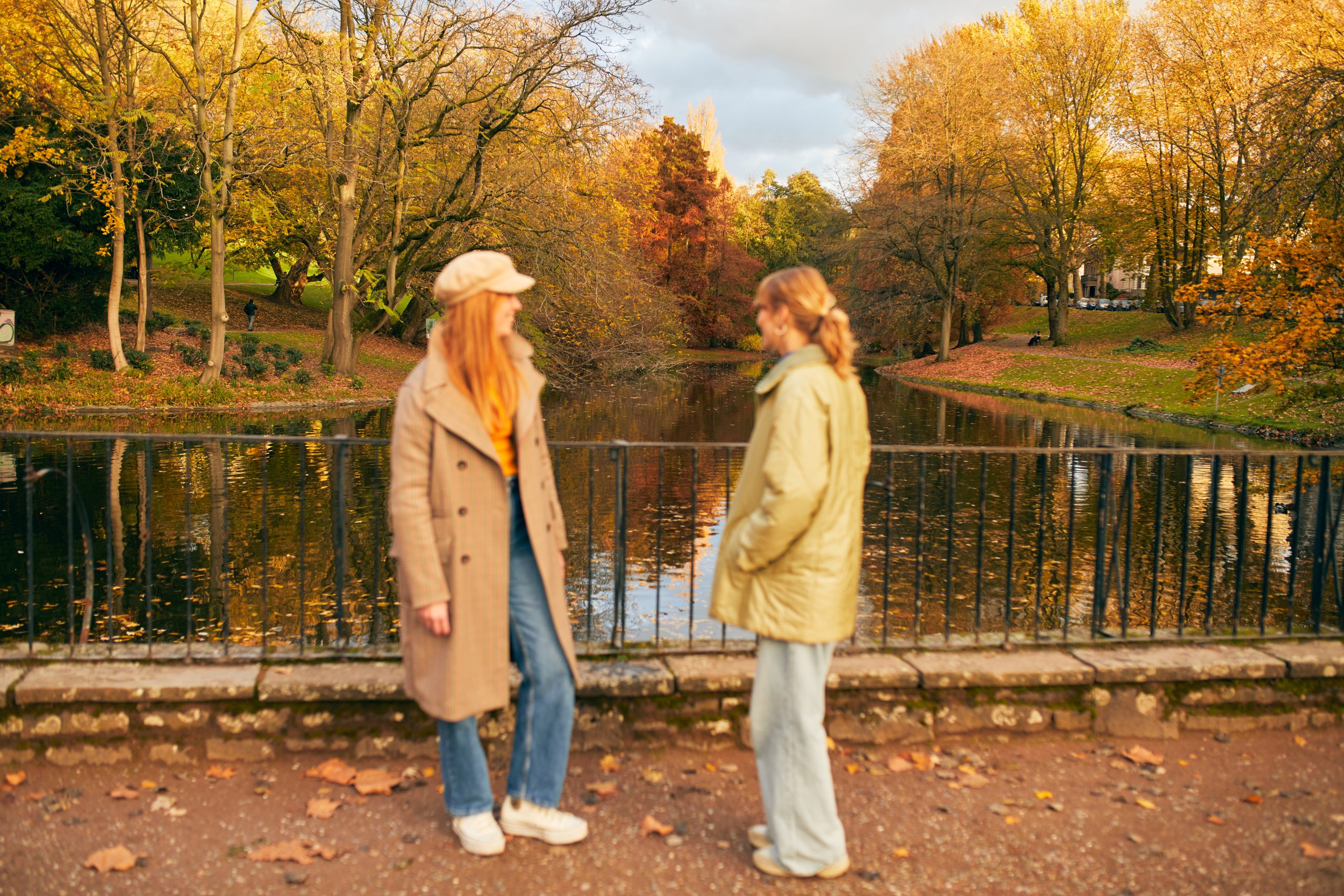 Zwei Menschen in Herbstmänteln stehen an einem schwarzen Metallzaun mit Blick auf einen Teich, umgeben von bunten Herbstbäumen mit orangefarbenen und gelben Blättern, die den Boden bedecken.