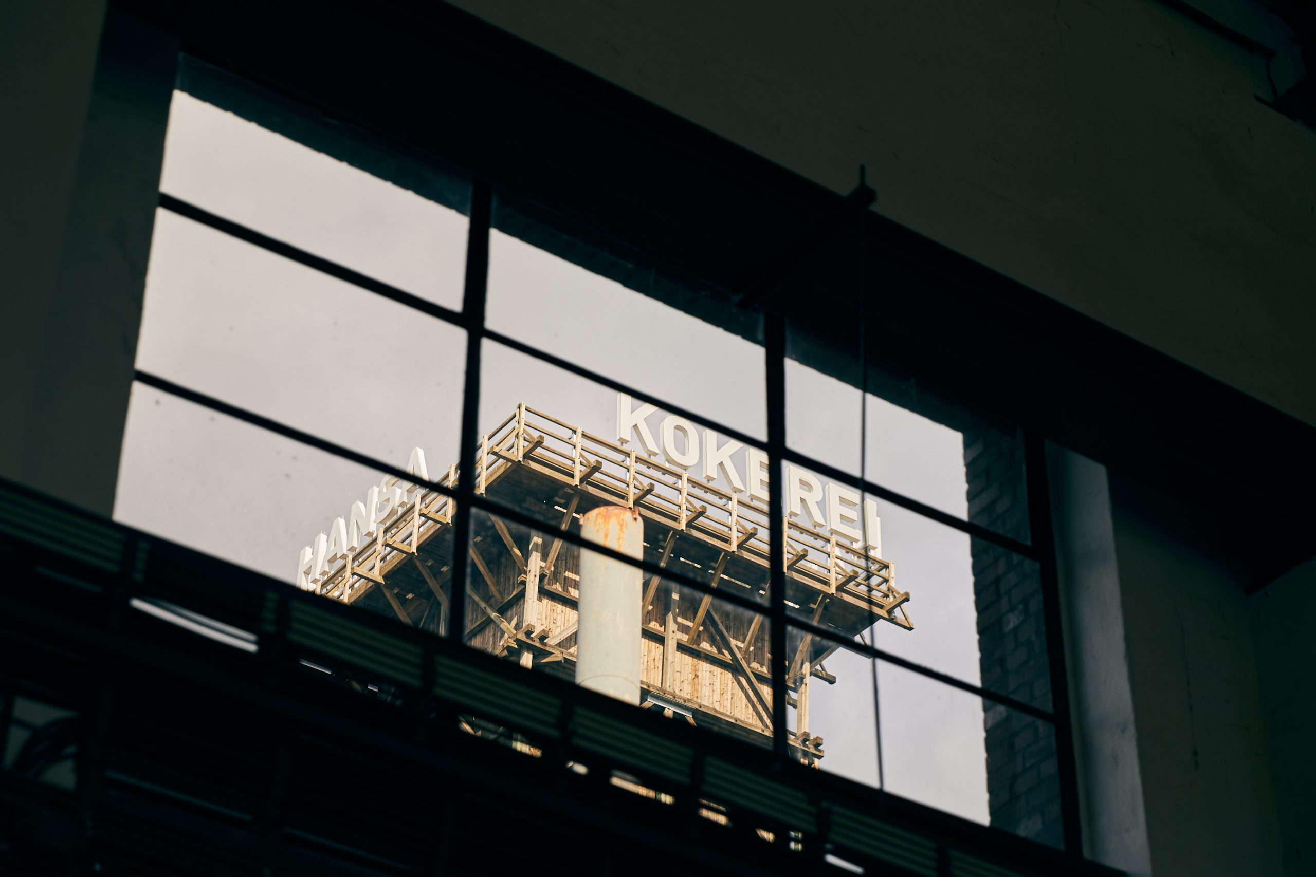 Ein hohes Industriegebäude mit dem teilweise sichtbaren Schriftzug KOKEREI ist durch große, gitterartige Fensterscheiben zu sehen, mit einigen Teilen eines dunklen Gebäuderahmens im Vordergrund.