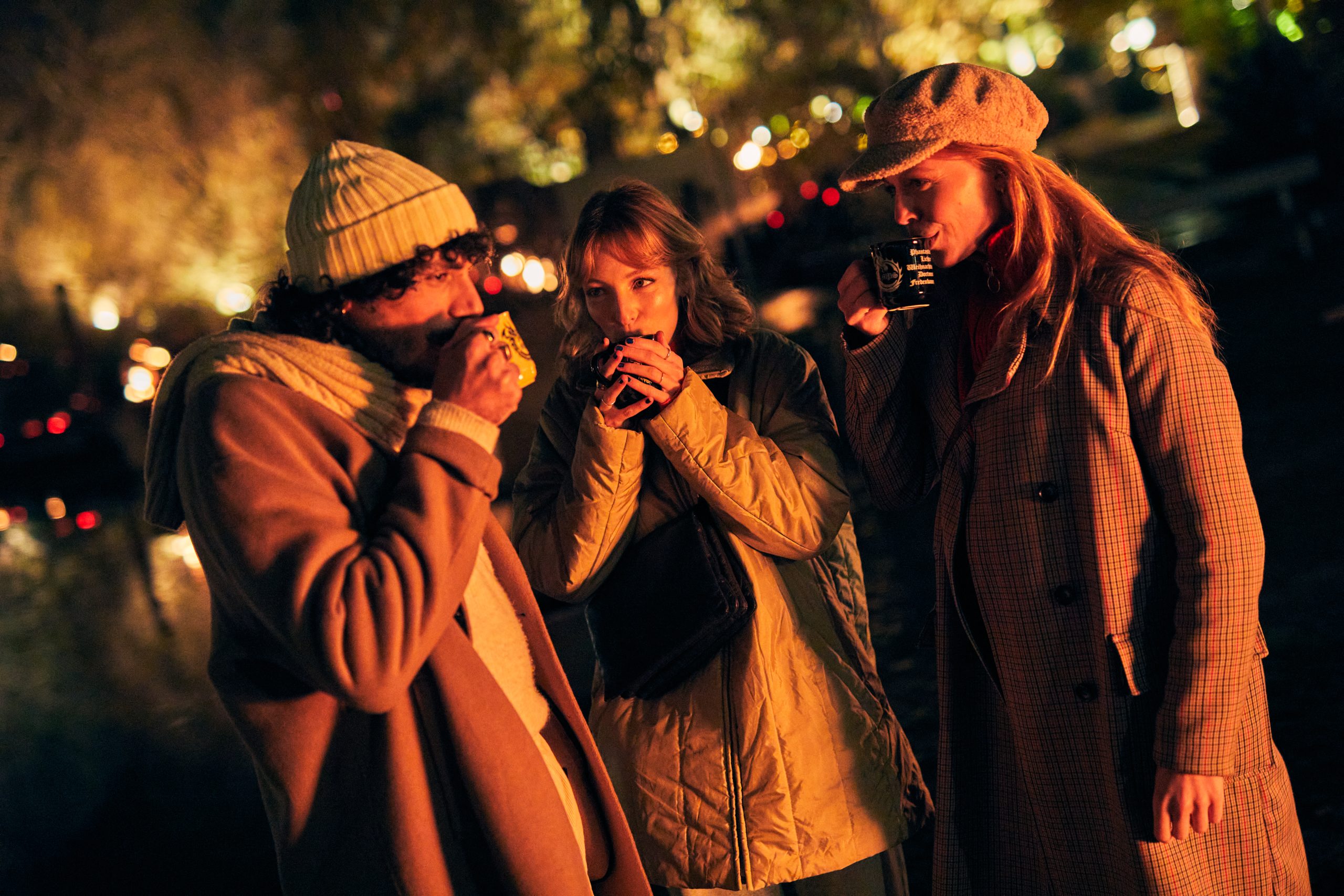 Drei in Mäntel und Hüte gehüllte Personen stehen nachts im Freien dicht beieinander und trinken aus einer Tasse. Warme Lichter im Hintergrund schaffen eine gemütliche, festliche Atmosphäre.