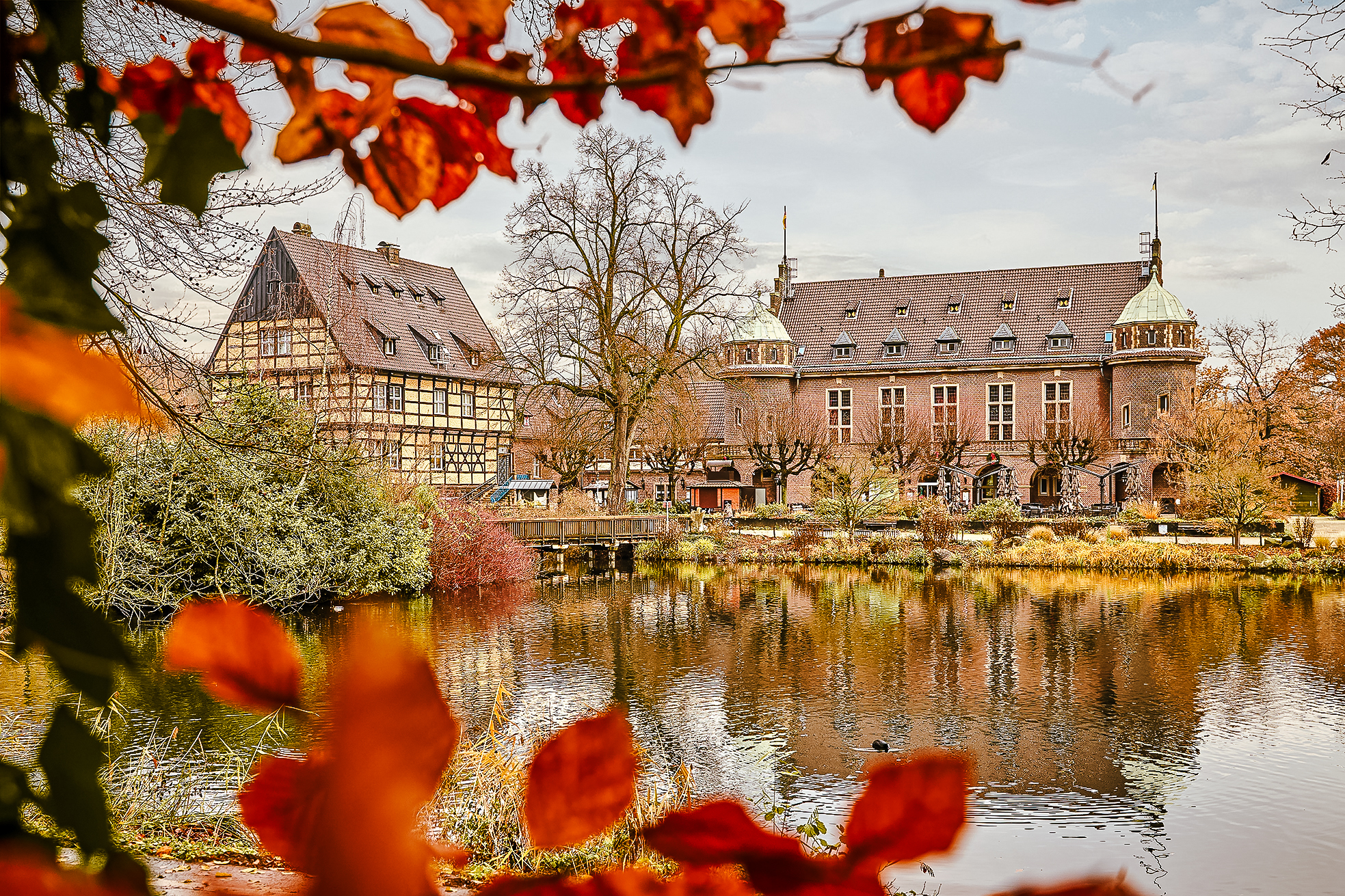 Ein malerischer Blick auf historische Gebäude an einem ruhigen Teich, umrahmt von Herbstlaub. Unter einem bewölkten Himmel spiegeln sich die Gebäude und Bäume im Wasser.
