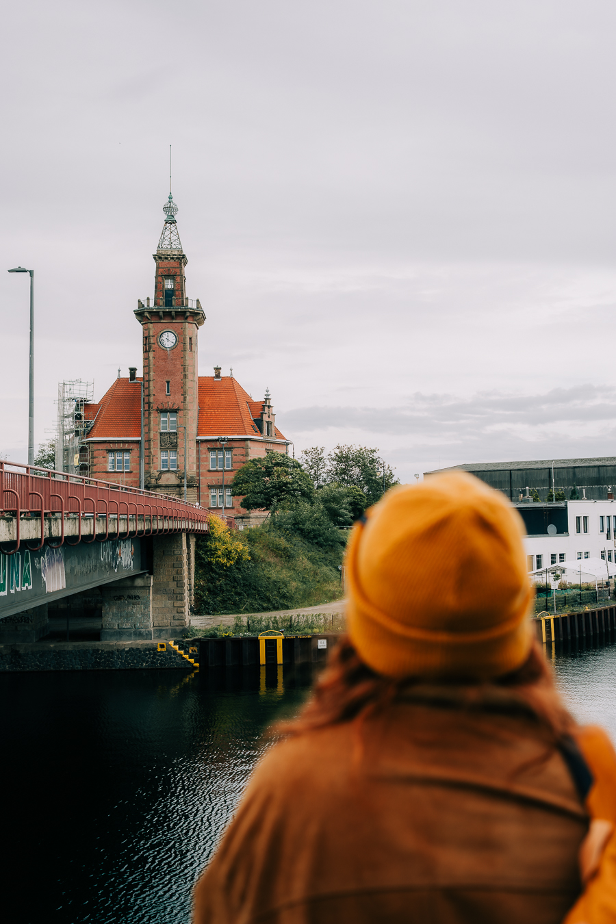 Eine Person mit gelbem Hut und Mantel steht am Wasser und blickt auf ein altes Backsteingebäude mit Uhrenturm und rotem Dach in der Nähe einer Brücke an einem bewölkten Tag.