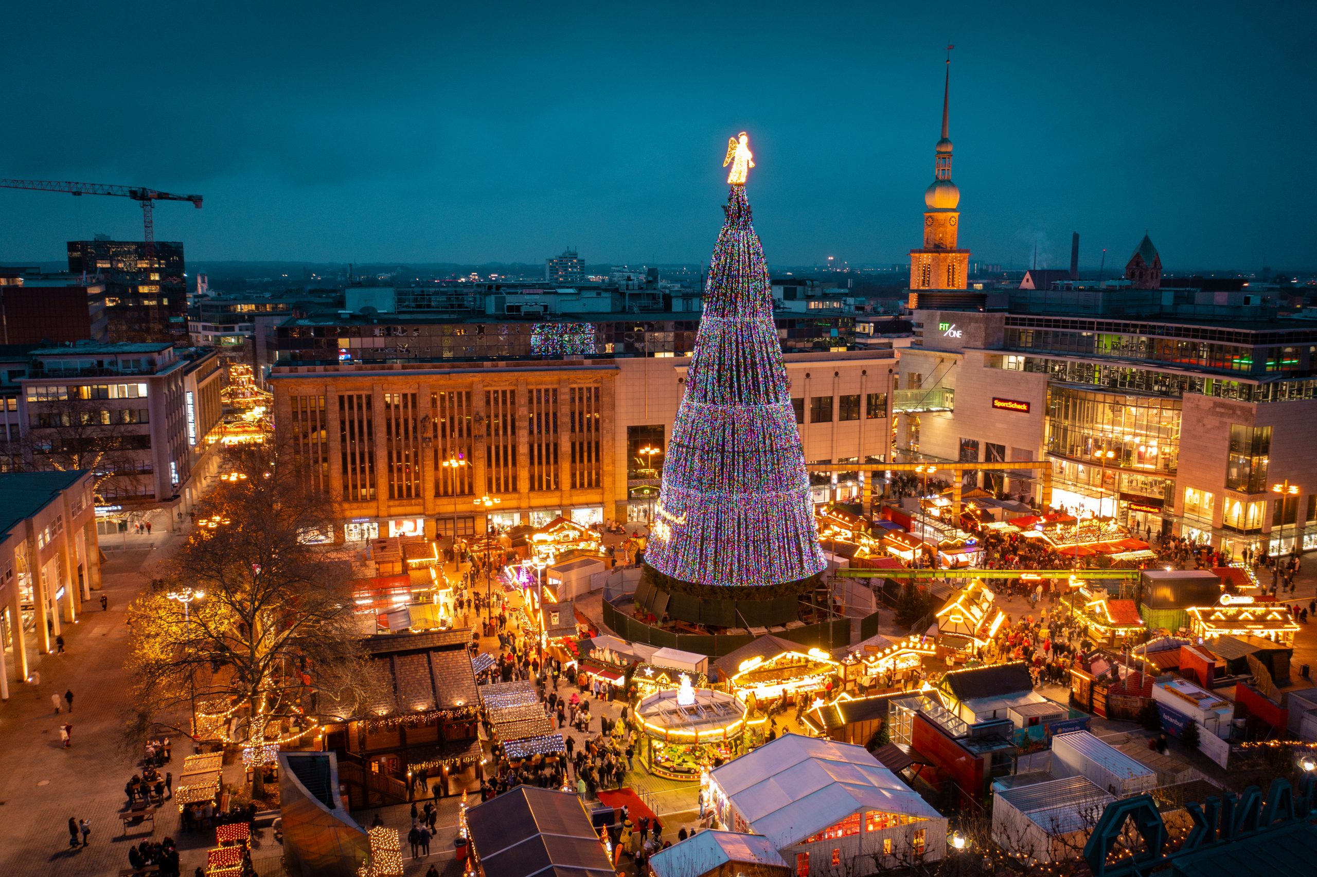 Ein festlicher Weihnachtsmarkt bei Nacht mit einem hohen, hell erleuchteten Weihnachtsbaum in der Mitte, umgeben von geschmückten Ständen, Menschen und städtischen Gebäuden unter einem dunkelblauen Himmel.