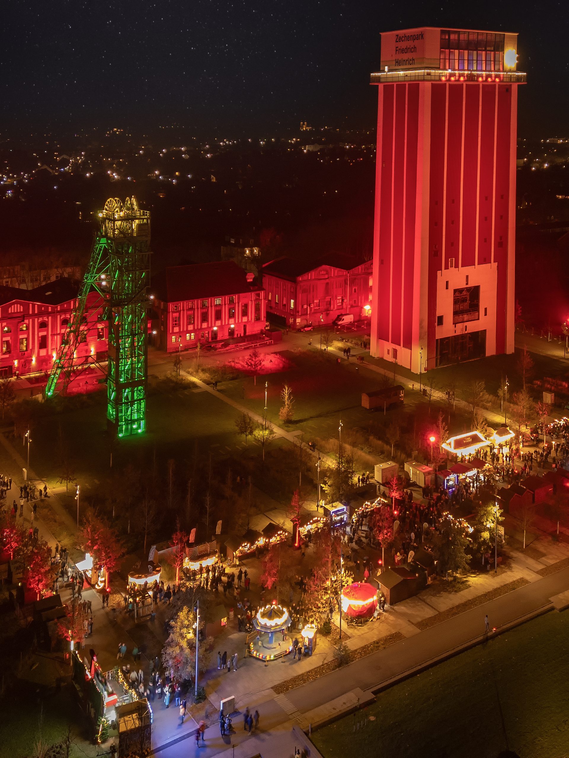 Eine nächtliche Luftaufnahme eines hell erleuchteten Festivals mit bunten Lichtern, Fahrgeschäften und Menschenmassen vor hohen beleuchteten Industriegebäuden und einem grün beleuchteten Turm mit einem Sternenhimmel darüber.