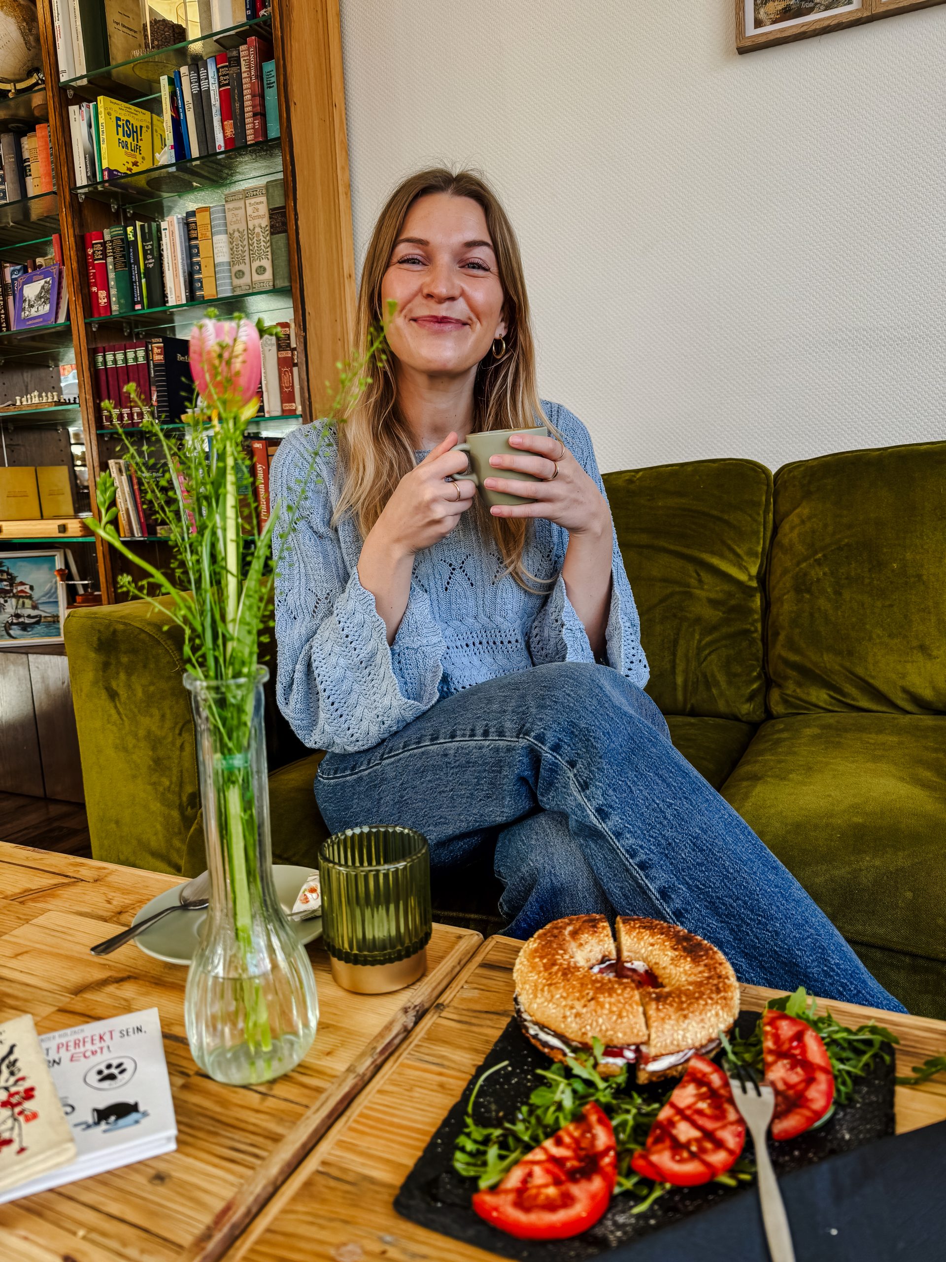 Eine Frau sitzt auf einer grünen Couch, lächelt und hält eine Tasse in der Hand. Vor ihr auf dem Holztisch stehen eine Vase mit Blumen, ein grünes Glas, ein Buch und ein Teller mit einem Bagel-Sandwich und geschnittenen Tomaten.