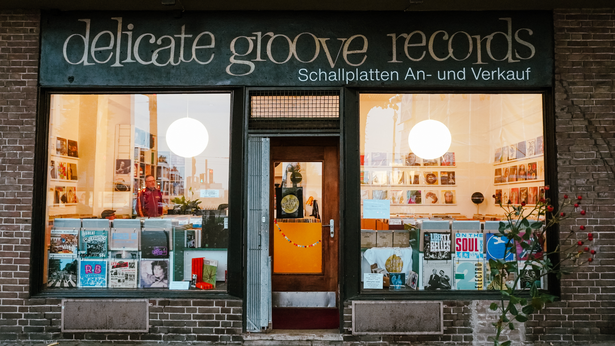 Ein Schallplattengeschäft mit großen Schaufenstern stellt Platten aus und hat darüber ein Schild mit der Aufschrift "delicate groove records Schallplatten An- und Verkauf". Die Eingangstür befindet sich in der Mitte zwischen den Fenstern.