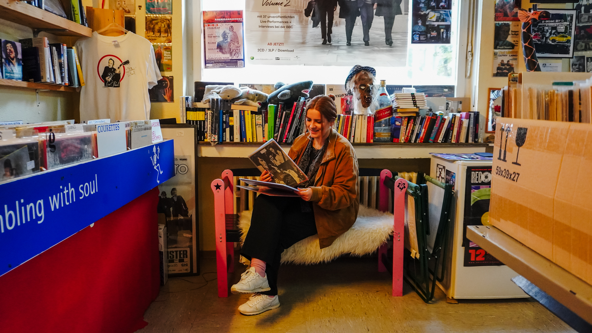 Eine Frau sitzt auf einer Bank in einem gemütlichen Plattenladen und lächelt, während sie eine Schallplatte betrachtet. Sie ist umgeben von Regalen mit Büchern, Schallplatten und Postern, und hinter ihr fällt warmes Licht durch ein Fenster.