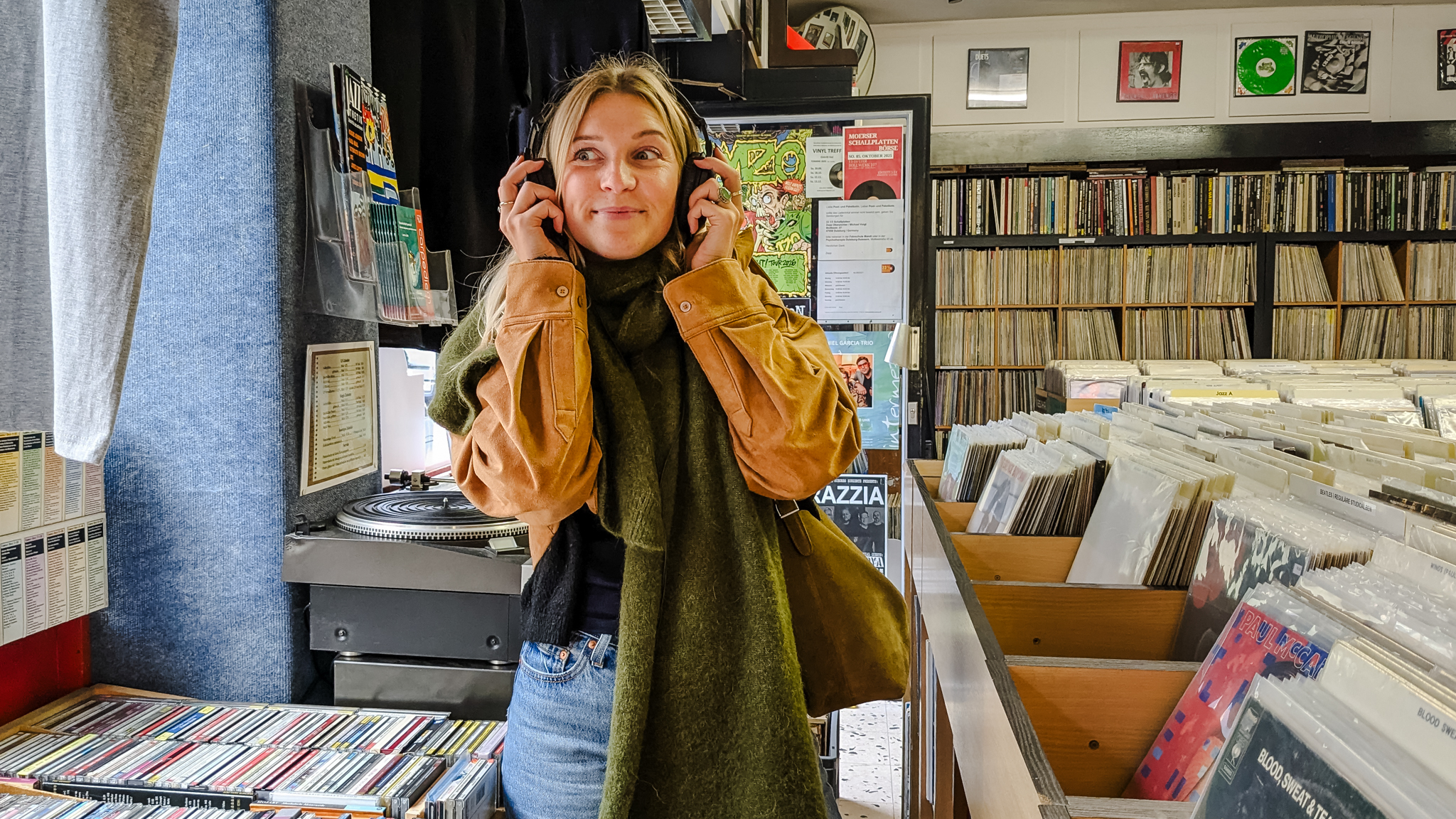 Eine junge Frau mit grünem Schal und brauner Jacke steht in einem Plattenladen und lächelt, während sie über Kopfhörer Musik hört. Sie ist umgeben von Schallplatten und Plattencovern in einem gemütlichen, bunten Laden.