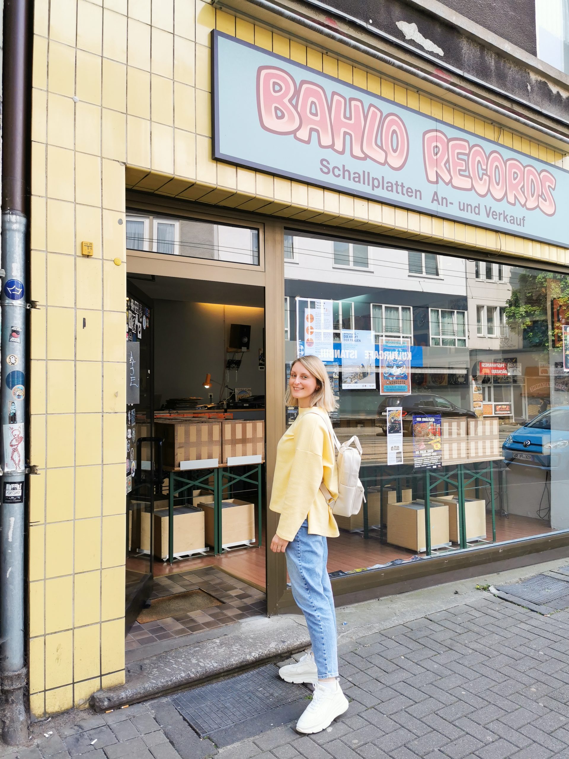 Eine Frau mit blondem Haar, die einen gelben Pullover, hellblaue Jeans und weiße Schuhe trägt, steht lächelnd am Eingang von Bahlo Records, einem Plattenladen mit gelben Kacheln an einer Stadtstraße.