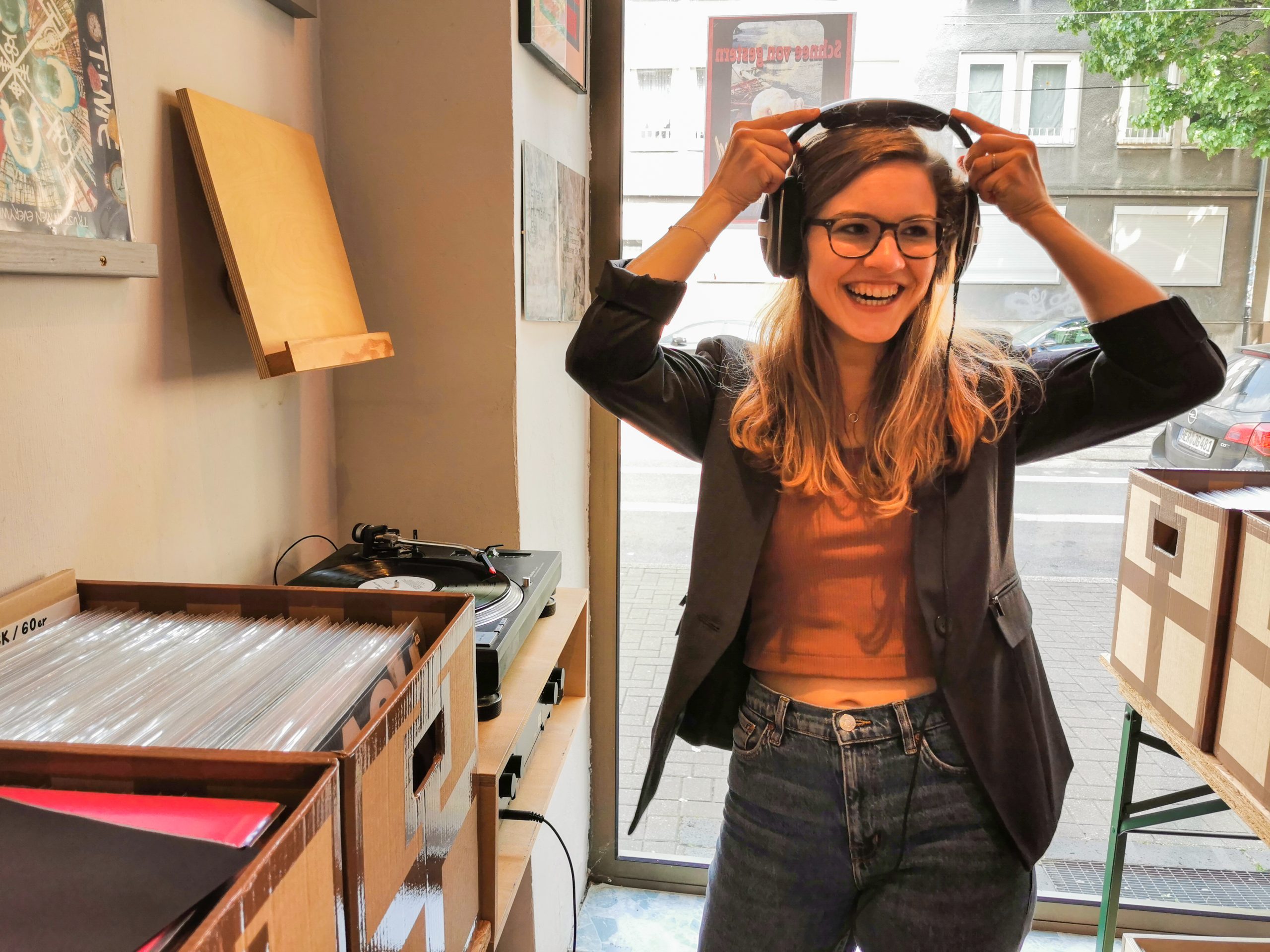 Eine lächelnde Frau mit Brille und Kopfhörern steht in einem Plattenladen neben Regalen mit Schallplatten, einem Plattenspieler und einem großen Schaufenster, aus dem man die geparkten Autos draußen sieht.