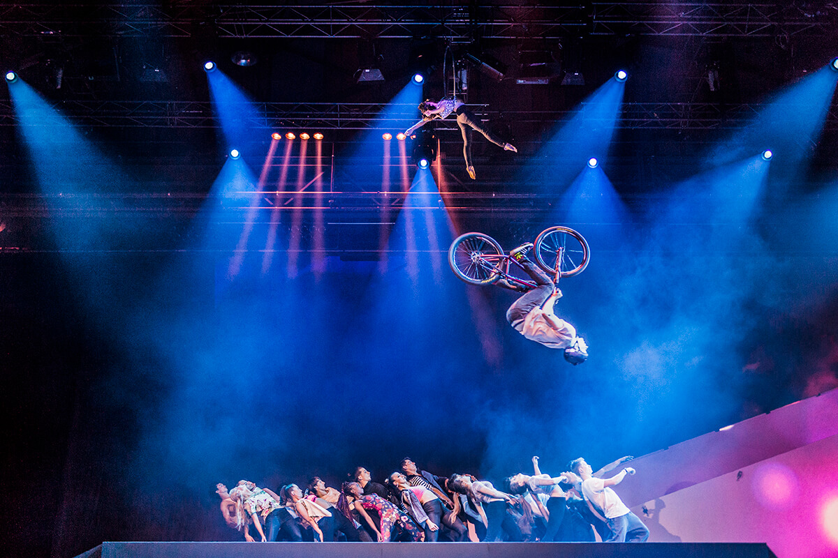 Das Bild zeigt eine Gruppe Tänzer und darüber einen BMXler, der einen Salto macht.
