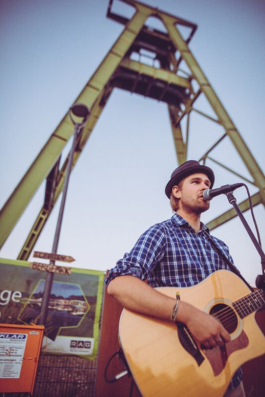 Das Bild zeigt einen Künstler mit Gitarre vor einem Fördergerüst.