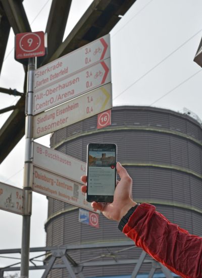 Das Foto zeigt einen Radfahrer vorm Gasometer Oberhausen, der den digitalen radtourenplaner.ruhr auf seinem Handy bedient