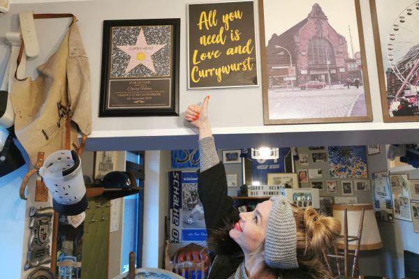 Das Foto zeigt Sandra vor einem Bild mit der Message "All you need is Love and Currywurst" beim Curry Heinz in Gelsenkirchen