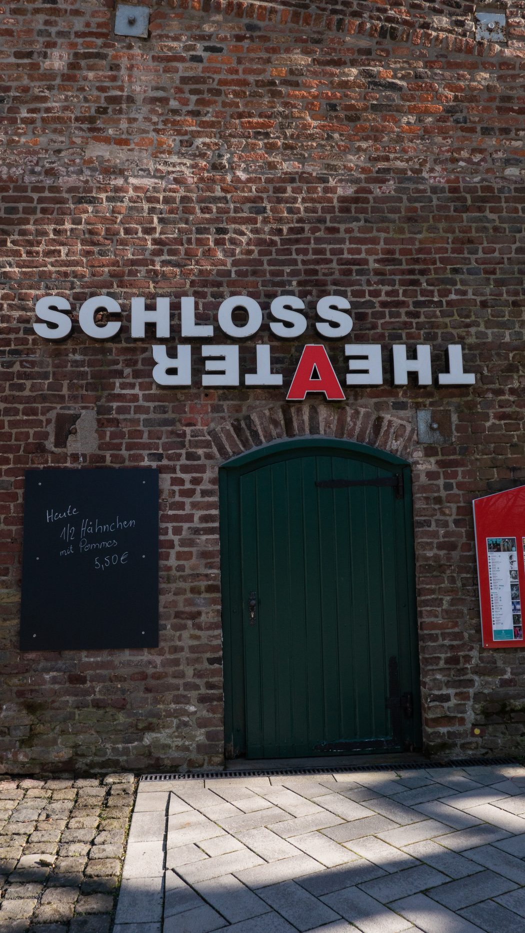 Eine grüne Rundbogentür in einer Backsteinmauer unter einem umgedrehten Schild mit der Aufschrift SCHLOSS THEATER. Auf der linken Seite befindet sich eine Tafel mit der Speisekarte in deutscher Sprache, auf der rechten Seite ein rot umrahmtes schwarzes Brett.