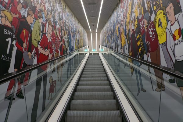 Eine breite Rolltreppe führt zwischen Wänden hinauf, die mit farbenfrohen Wandgemälden bedeckt sind, auf denen Menschenmassen in Sporttrikots und Fankleidung abgebildet sind, was eine lebhafte Atmosphäre schafft.