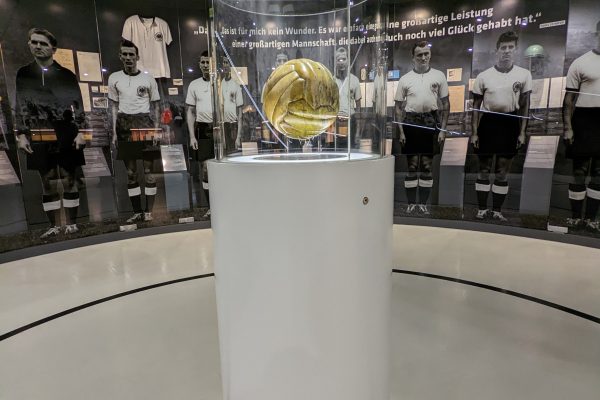 Ein alter goldener Fußball wird in einer Vitrine auf einem weißen Sockel ausgestellt, umgeben von Schwarz-Weiß-Fotos historischer Fußballspieler und Mannschaftserinnerungsstücken an den Wänden dahinter.