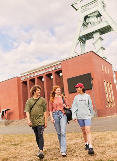 Drei junge Erwachsene spazieren und lächeln gemeinsam vor dem Deutschen Bergbau-Museum, einem roten Backsteinmuseum in Deutschland, mit einem Industrieturm im Hintergrund unter einem teilweise bewölkten Himmel.