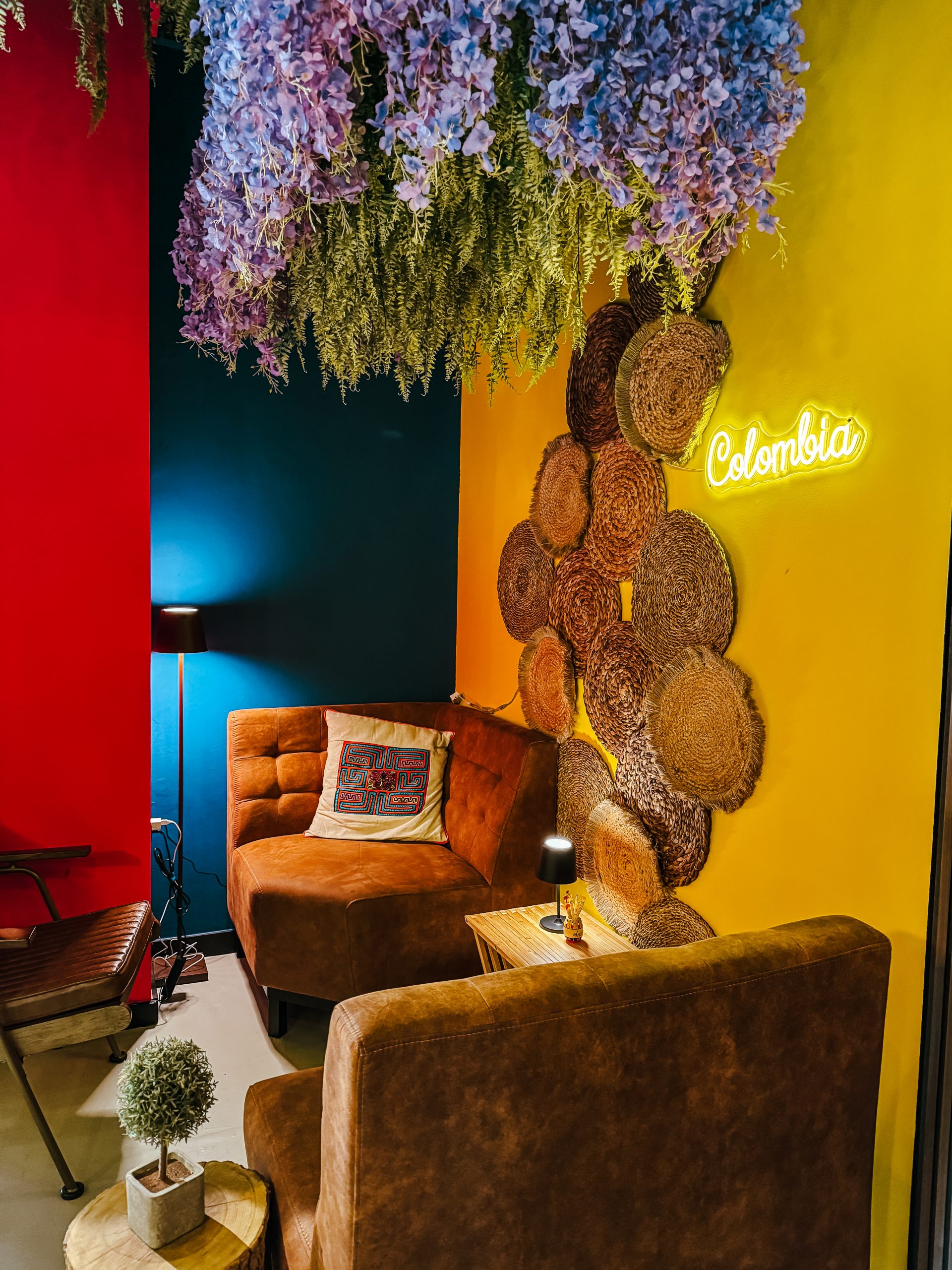 Gemütliche Sitzecke mit braunen Stühlen, kleinen Tischen und Kissen. Die Wände sind rot, blau und gelb und mit geflochtenen Körben und einem Neonschild "Kolumbien" dekoriert. Lila Blumen und Grünzeug hängen von der Decke.