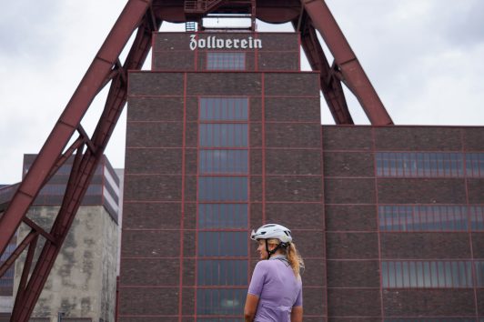 Ein Radfahrer mit Helm und Sportkleidung steht neben einem Fahrrad auf einer Wiese, im Hintergrund die rote Industrieanlage der Zeche Zollverein und Backsteinbauten.