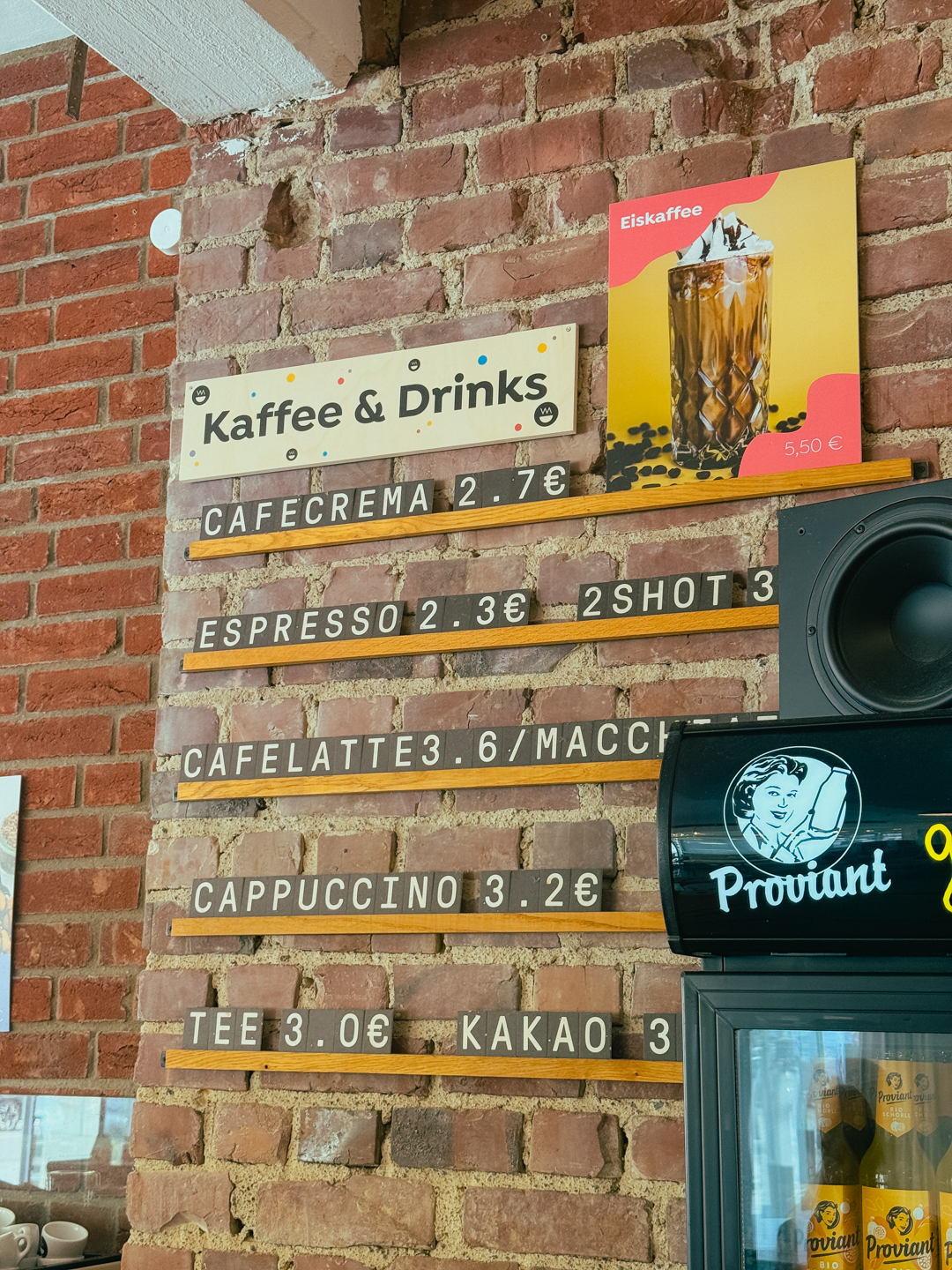 Speisekarte auf einer Backsteinmauer mit der Auflistung von Kaffee und Getränken mit Preisen für Cafecrema, Espresso, Cafelatte, Cappuccino, Tee und Kakao. Darüber ist ein Eiskaffee-Poster mit einem Bild des Getränks angebracht.