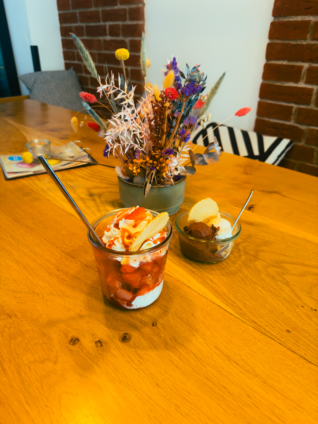 Ein Holztisch mit einem Glas Schlagsahne und einem Erdbeerdessert, eine Schale mit Eis und Waffelröllchen und eine Vase mit bunten Trockenblumen im Hintergrund.