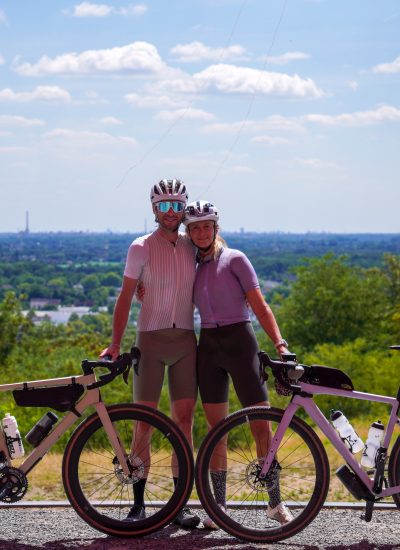 Ein lächelnder Mann und eine lächelnde Frau in Radfahrerkleidung stehen Seite an Seite mit ihren beladenen Fahrrädern und posieren auf einer Bergkuppe mit einer malerischen Landschaft und der entfernten Stadtsilhouette hinter ihnen unter einem teilweise bewölkten Himmel.