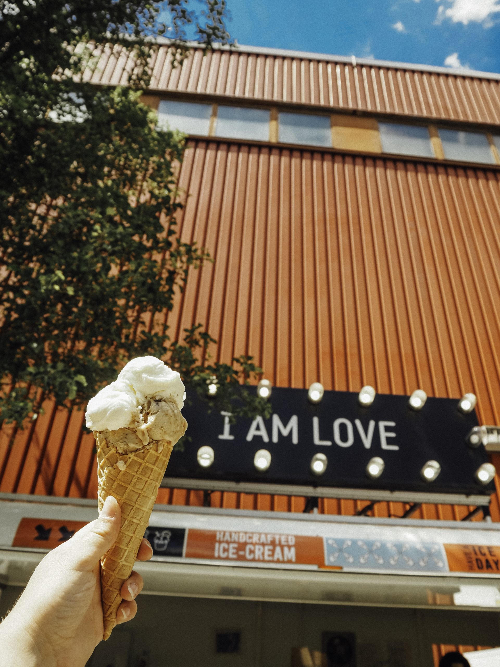 Eine Hand hält eine Eiswaffel mit zwei Kugeln vor einem roten Gebäude. Auf dem Gebäude befindet sich ein Schild mit der Aufschrift I AM LOVE in Leuchtbuchstaben, im Hintergrund sind Bäume und blauer Himmel zu sehen.