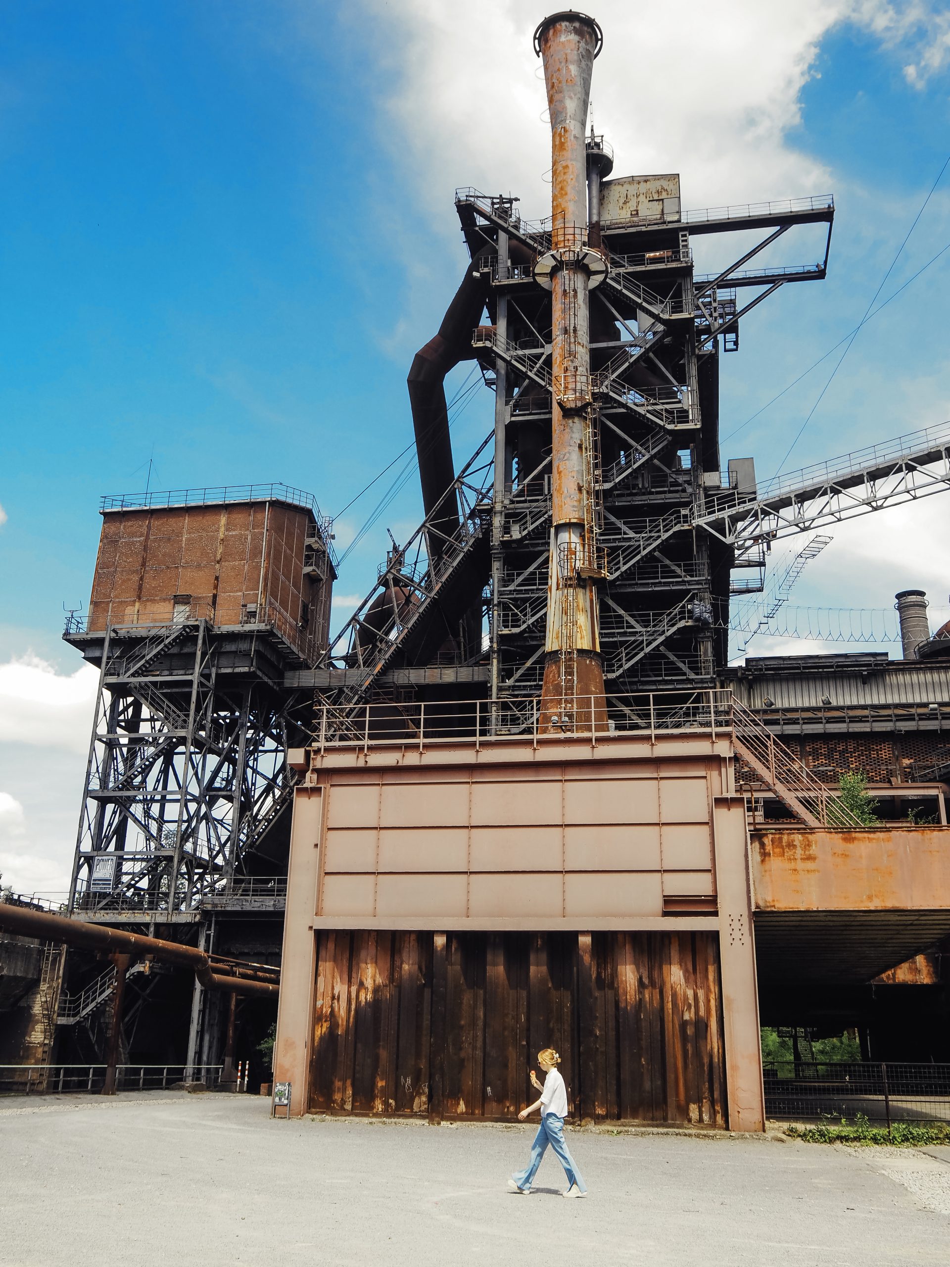 Eine leicht bekleidete Person geht unter einem teilweise bewölkten Himmel an einem großen, rostigen Industriegebäude mit hohen Schornsteinen und Metallgerüsten vorbei.