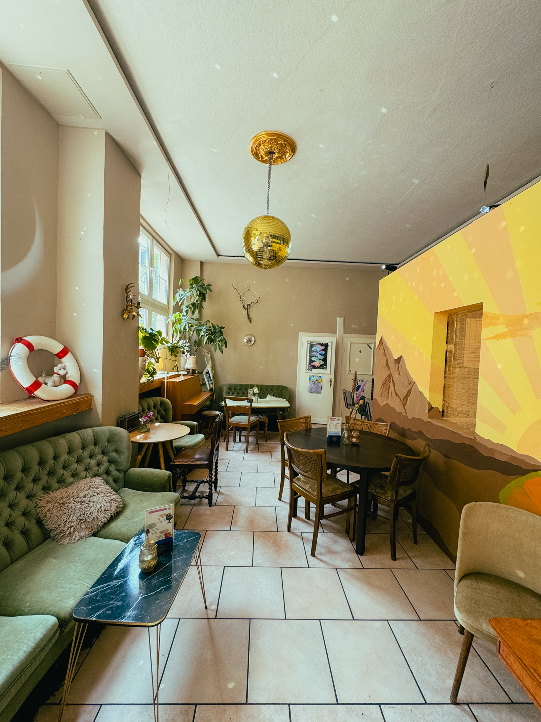 Ein gemütliches Café mit grünen und braunen Möbeln, einem Wandgemälde mit Bergen und einem gelben Sonnenstrahl an der Wand, einer goldenen Discokugel, die von der Decke hängt, und Pflanzen, die die Fensterbank schmücken.