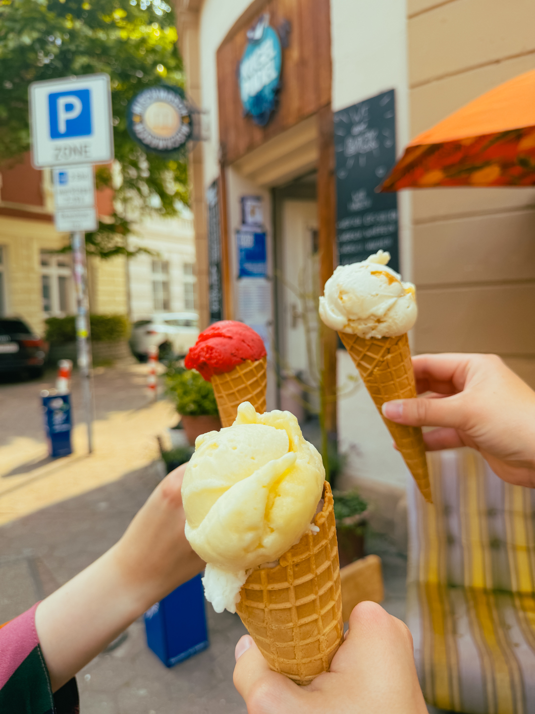Drei Personen halten Eistüten mit verschiedenen Geschmacksrichtungen vor einer Eisdiele an einem sonnigen Tag; im Hintergrund sind Straßenschilder, eine Tafel mit der Speisekarte und ein bunter Regenschirm zu sehen.