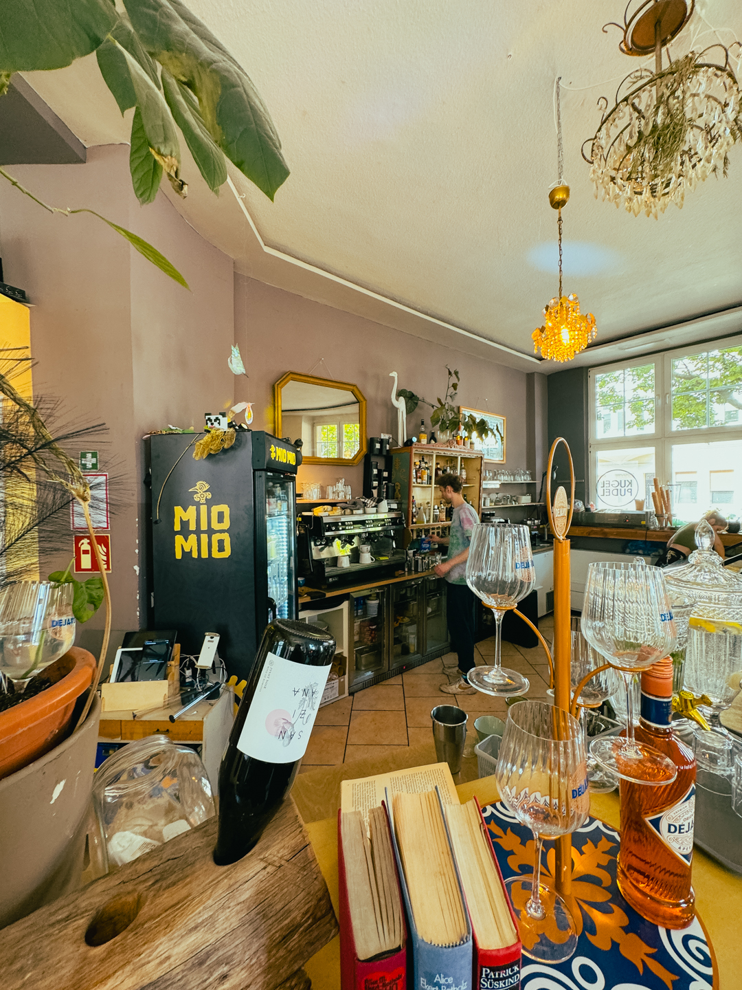 Ein gemütliches Café mit einem Tresen, Weinflaschen, Glaswaren, Topfpflanzen, Büchern und einem schwarzen Kühlschrank mit der Aufschrift MIO MIO. Durch große Fenster strömt Sonnenlicht, und von der Decke hängt ein Kronleuchter.