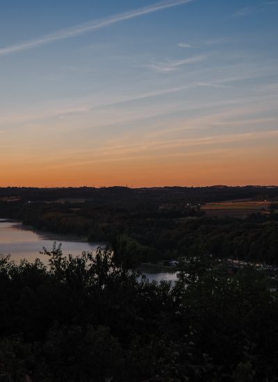 Ein ruhiger Fluss schlängelt sich durch eine üppige, grüne Landschaft bei Sonnenuntergang, mit orangefarbenen und blauen Farbtönen am Himmel und Silhouetten von Bäumen und Hügeln in der Ferne.