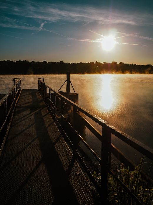 Ein Metallsteg mit Geländer führt über einen ruhigen See bei Sonnenaufgang, wenn Nebel vom Wasser aufsteigt und die Sonne über dem baumbestandenen Horizont scheint. Schatten erstrecken sich über den Steg.