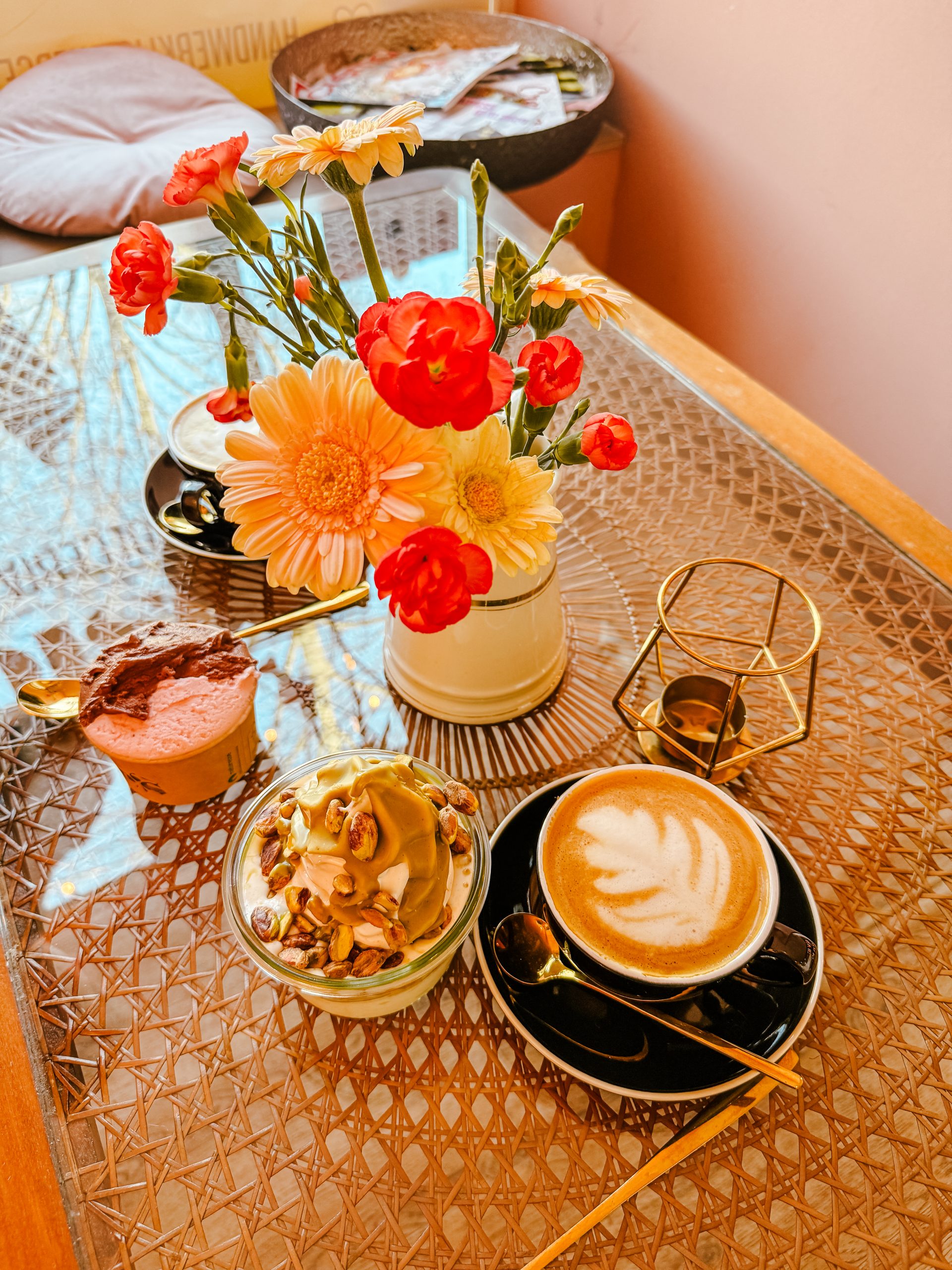 Ein Glastisch mit einem Milchkaffee, einer Glasschale mit Pistaziendessert, einem kleinen Schokoladenkuchen, einer Vase mit farbenfrohen Blumen und einem goldenen Kerzenständer, die ordentlich in einem gemütlichen Rahmen angeordnet sind.