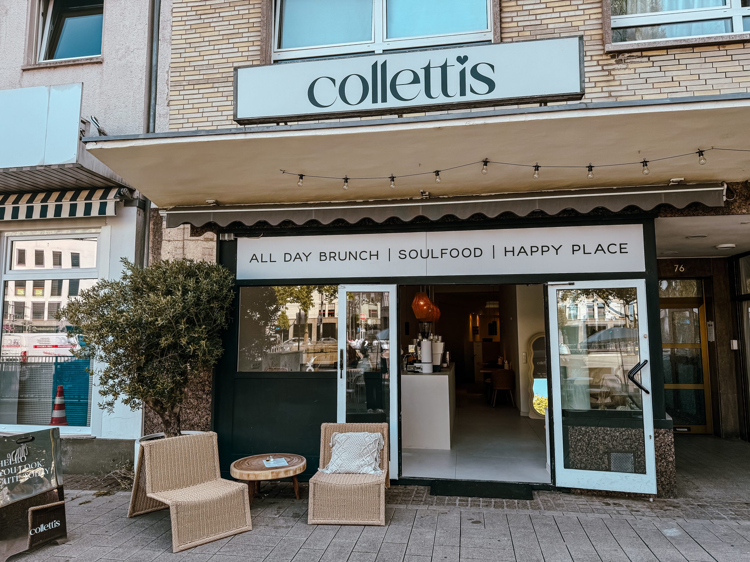 Straßenansicht eines Cafés namens collettis mit Korbstühlen und einem Tisch im Freien, großen Fenstern und einem Schild mit der Aufschrift All Day Brunch | Soulfood | Happy Place über dem Eingang.